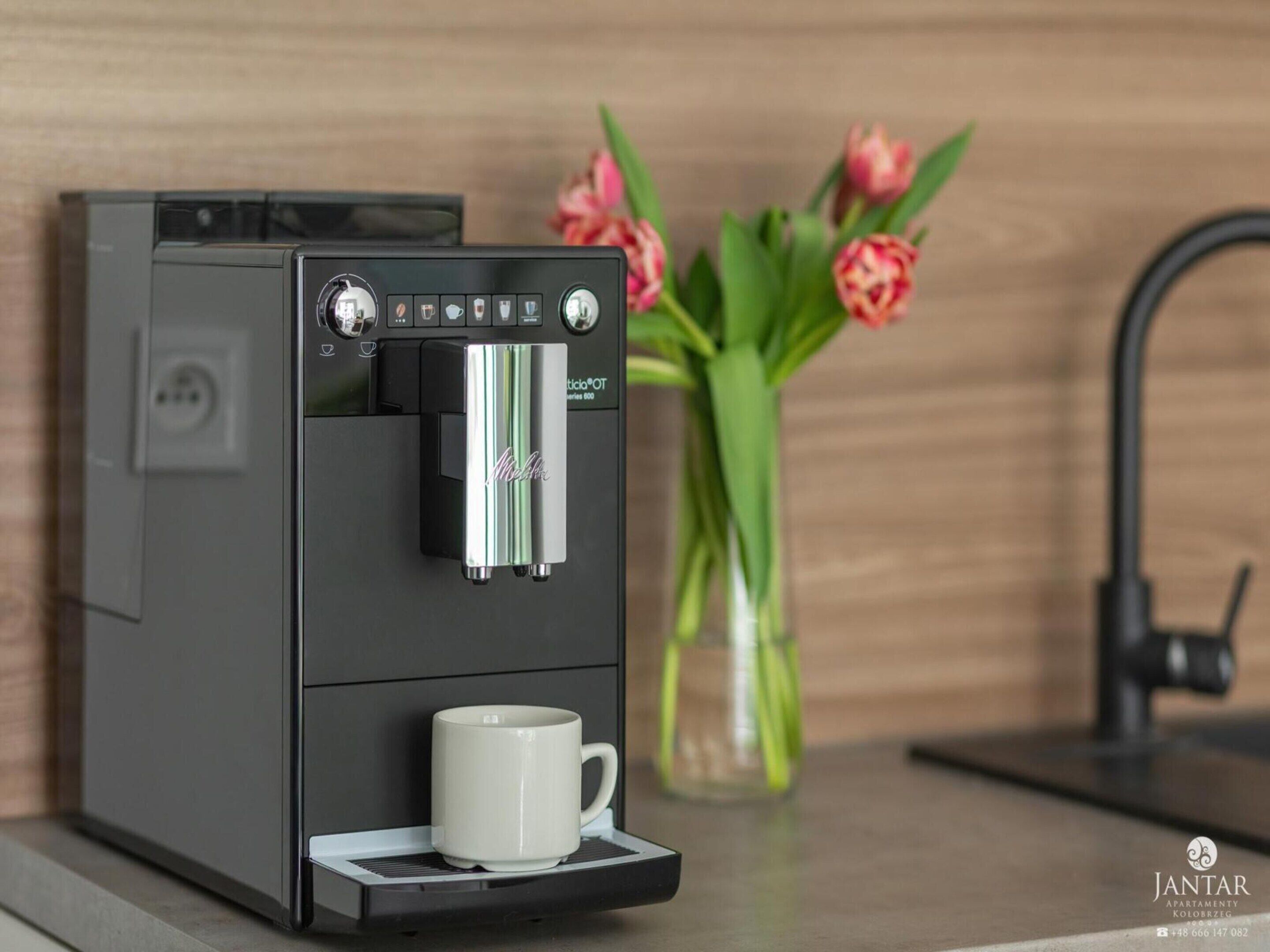 A coffee machine with a water reservoir and a cup holder, placed on a countertop with a vase of tulips in the background.