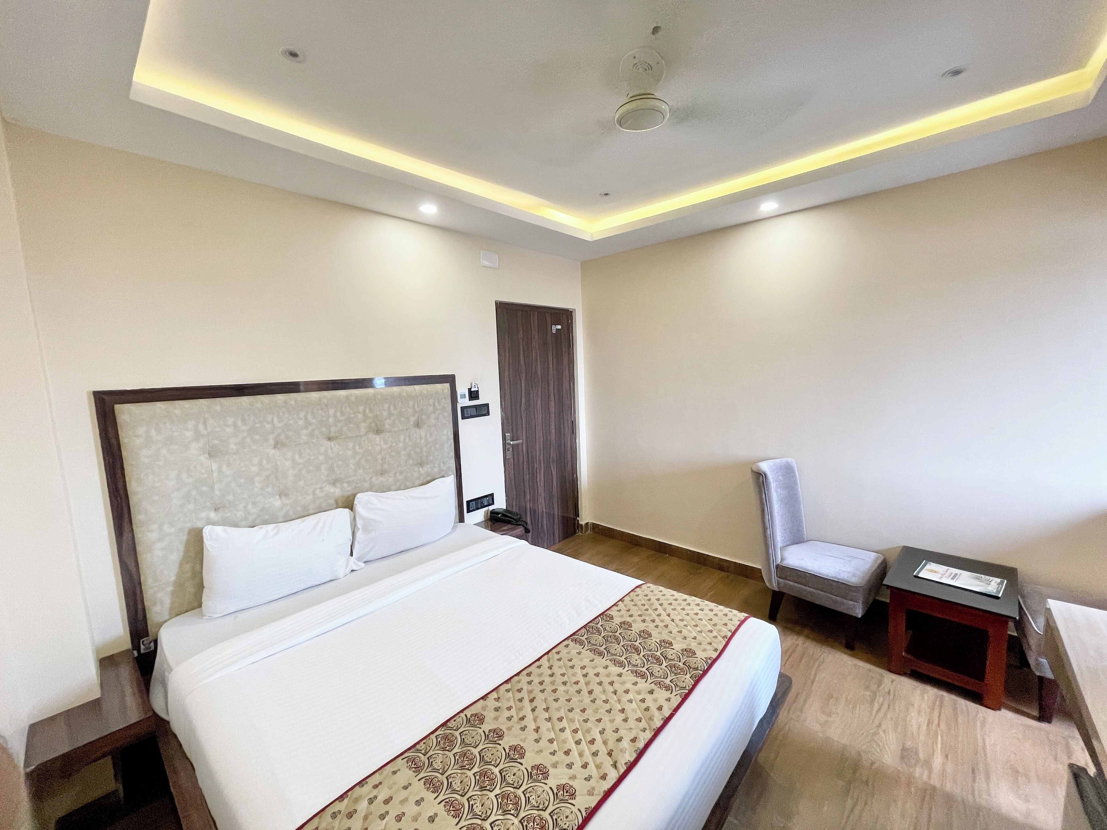Quarto de hotel com uma cama grande, uma cadeira, uma mesa pequena e uma porta.
