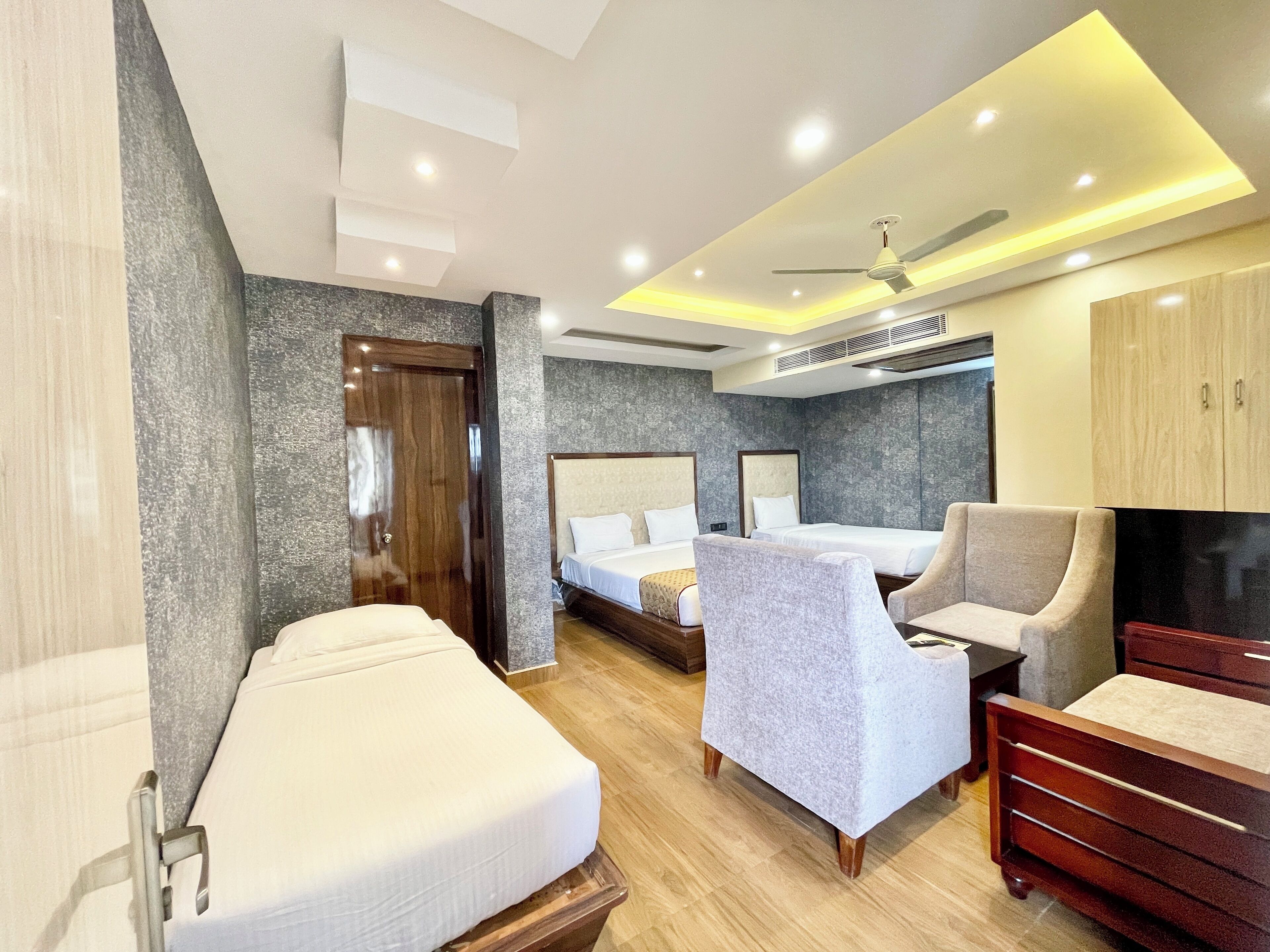 A hotel room with two beds, a sofa, a chair, a wardrobe, and a ceiling fan.