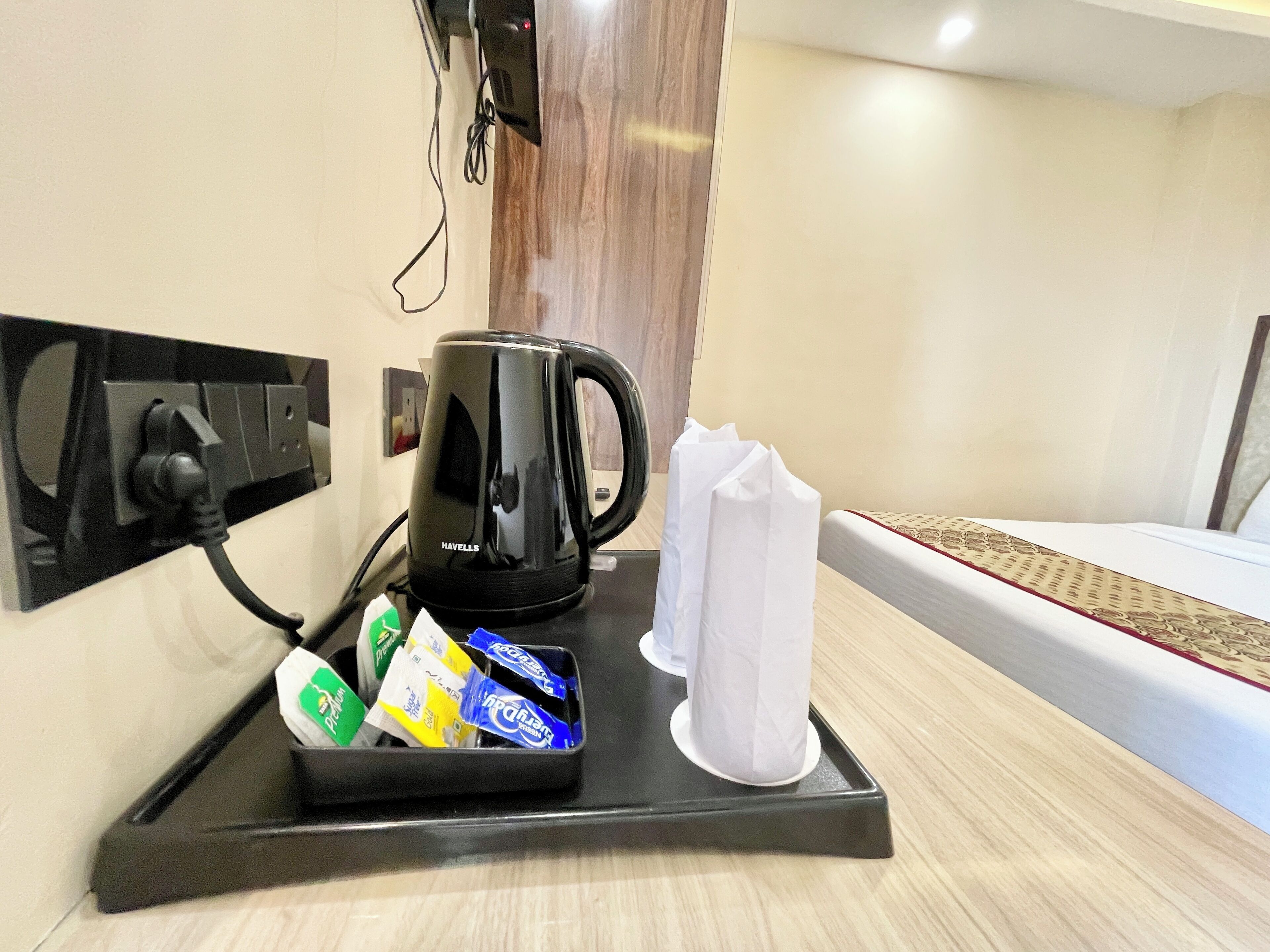 Eine Kaffeemaschine auf einem Tablett mit Einwegbechern und Kaffeekapseln, eine Steckdose und ein Bett mit einem gemusterten Bettbezug.