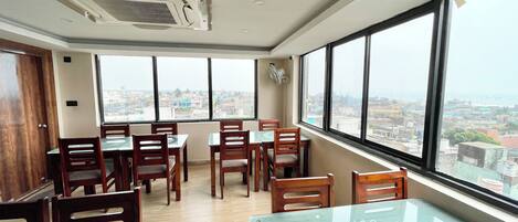 A dining area with tables and chairs, large windows with a city view, and an air conditioning unit on the ceiling.