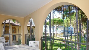 A balcony with a table and chairs, overlooking a green area with trees and a building in the background.