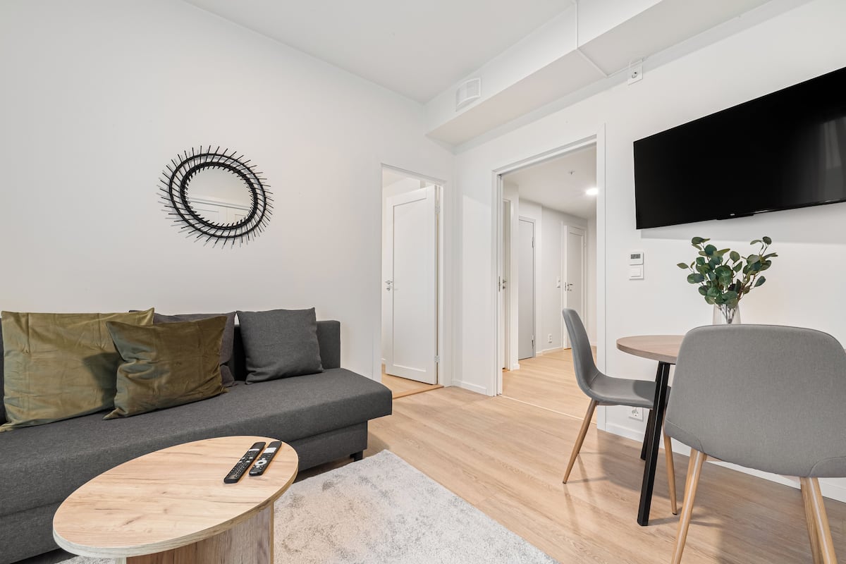 A modern living room with a grey sofa, a round coffee table, a TV mounted on the wall, and a dining area with a table and chairs.