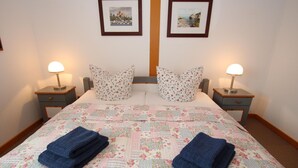 A neatly made bed with floral bedding, two pillows, and folded towels. There are two bedside tables with lamps and framed pictures on the wall above. - Appartementhaus auf Pellworm (Pellworm)
