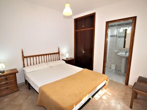 Een slaapkamer met een bed, nachtkastje, lamp, houten garderobe en een badkamer door een geopende deur.