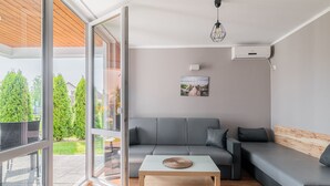 A modern living room with a grey sofa, a wooden coffee table, and a large window offering a view of greenery and a bridge. - Holiday Homes With Pool, Close to the Beach (Ustronie Morskie)