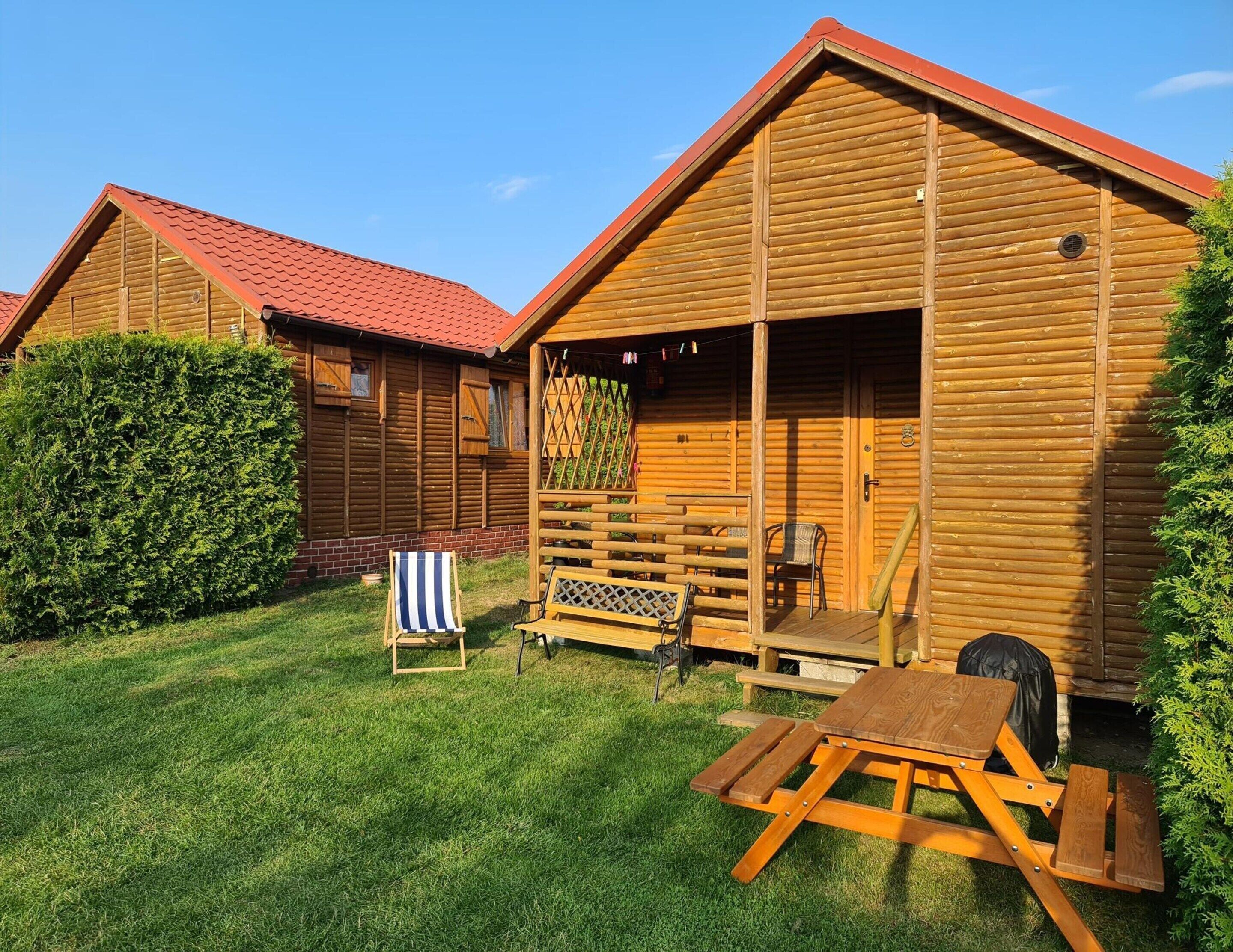 Une cabane en bois avec un toit rouge, un espace patio avec une table et des chaises, et une haie soigneusement taillée.