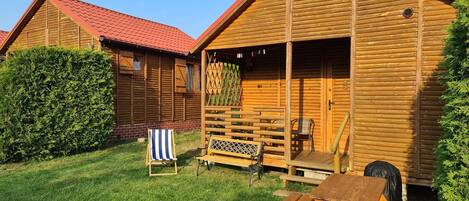 A wooden cabin with a red roof, a patio area with a table and chairs, and a neatly trimmed hedge.