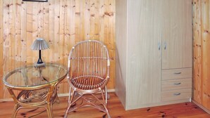Une pièce avec un mur lambrissé en bois, une table à plateau en verre, une chaise en osier et une armoire à deux portes et trois tiroirs.