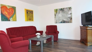 Un salon moderne avec un canapé rouge, un fauteuil rouge et une table basse en verre. Un téléviseur à écran plat est posé sur un meuble en bois, et trois tableaux abstraits ornent les murs.