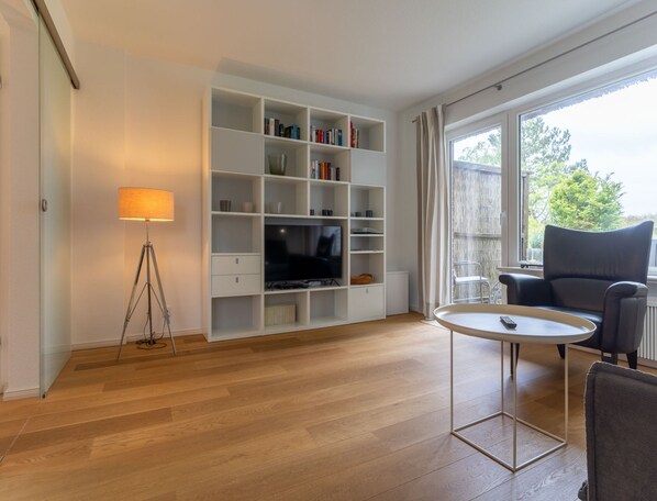 A modern living room with a wooden floor, a white entertainment unit with books and decorative items, a floor lamp, a round coffee table, and a grey sofa.