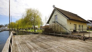 Een houten steiger die uitloopt in een rustig wateroppervlak, een klein gebouw met een schuin dak en een schotelantenne, en een rij bomen met groene bladeren.