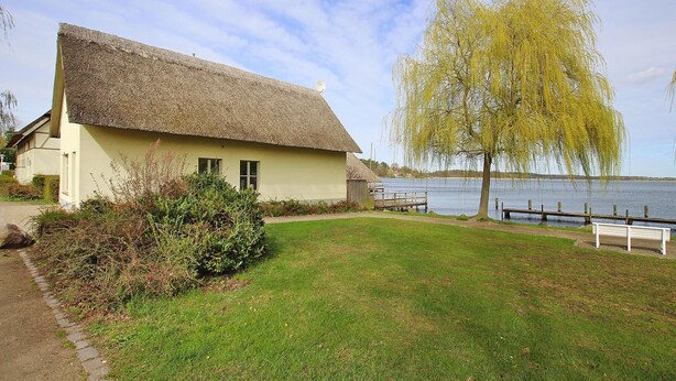 Een rieten huisje aan een meer, met een bankje en een treurwilg.