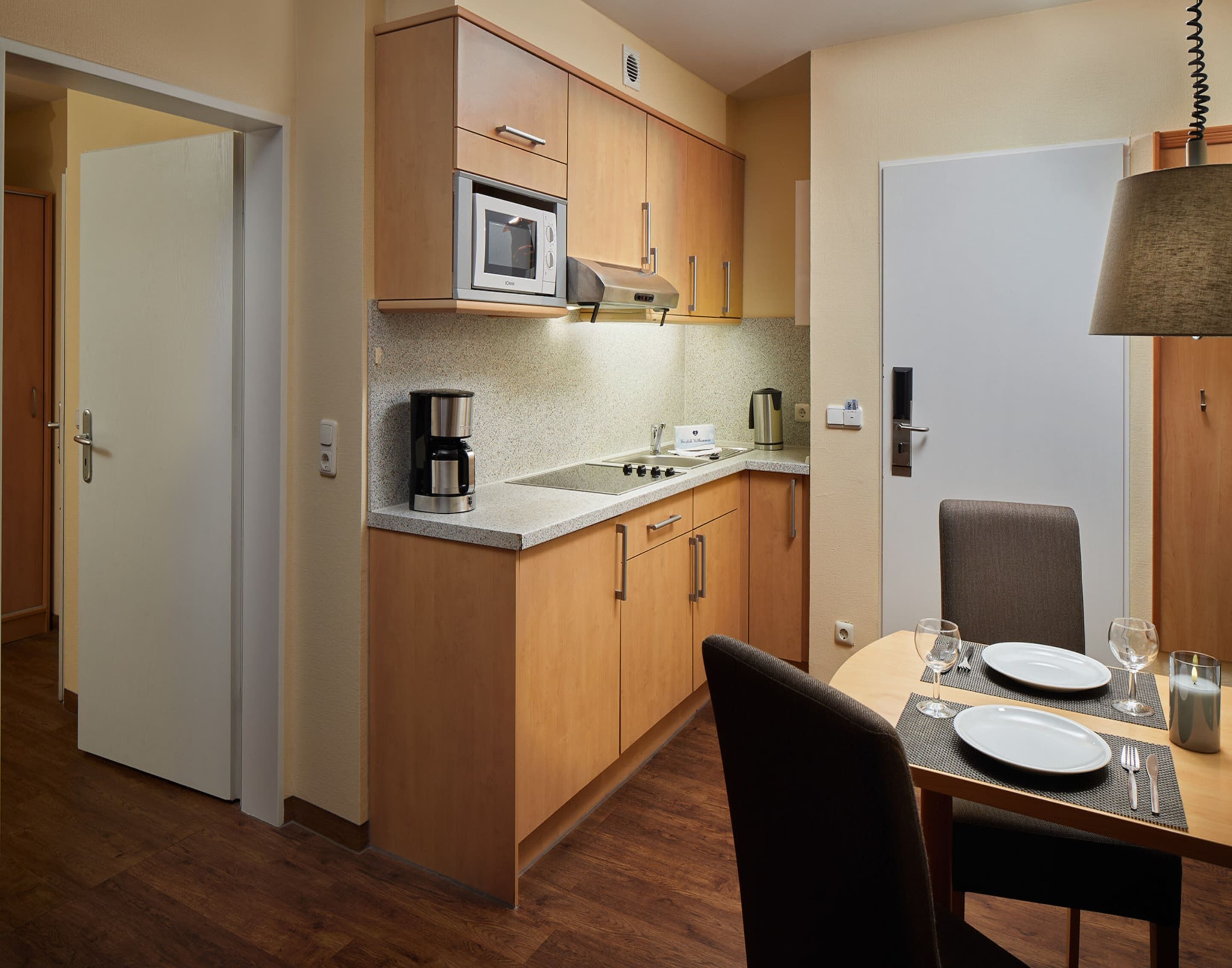 A compact kitchen with wooden cabinets, a built-in microwave, a coffee maker, and a sink. There is a dining table with chairs and a candle on it.