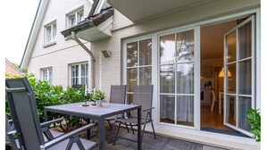 Un patio con una mesa y sillas, un ventanal corredero que da a una habitación y una pequeña zona ajardinada.