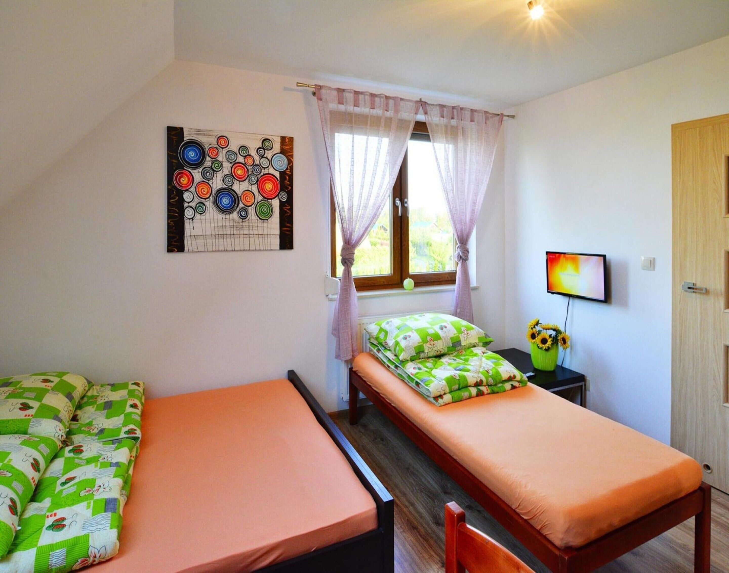 A small, well-lit room with two single beds, a window with curtains, a wall-mounted TV, and a painting.