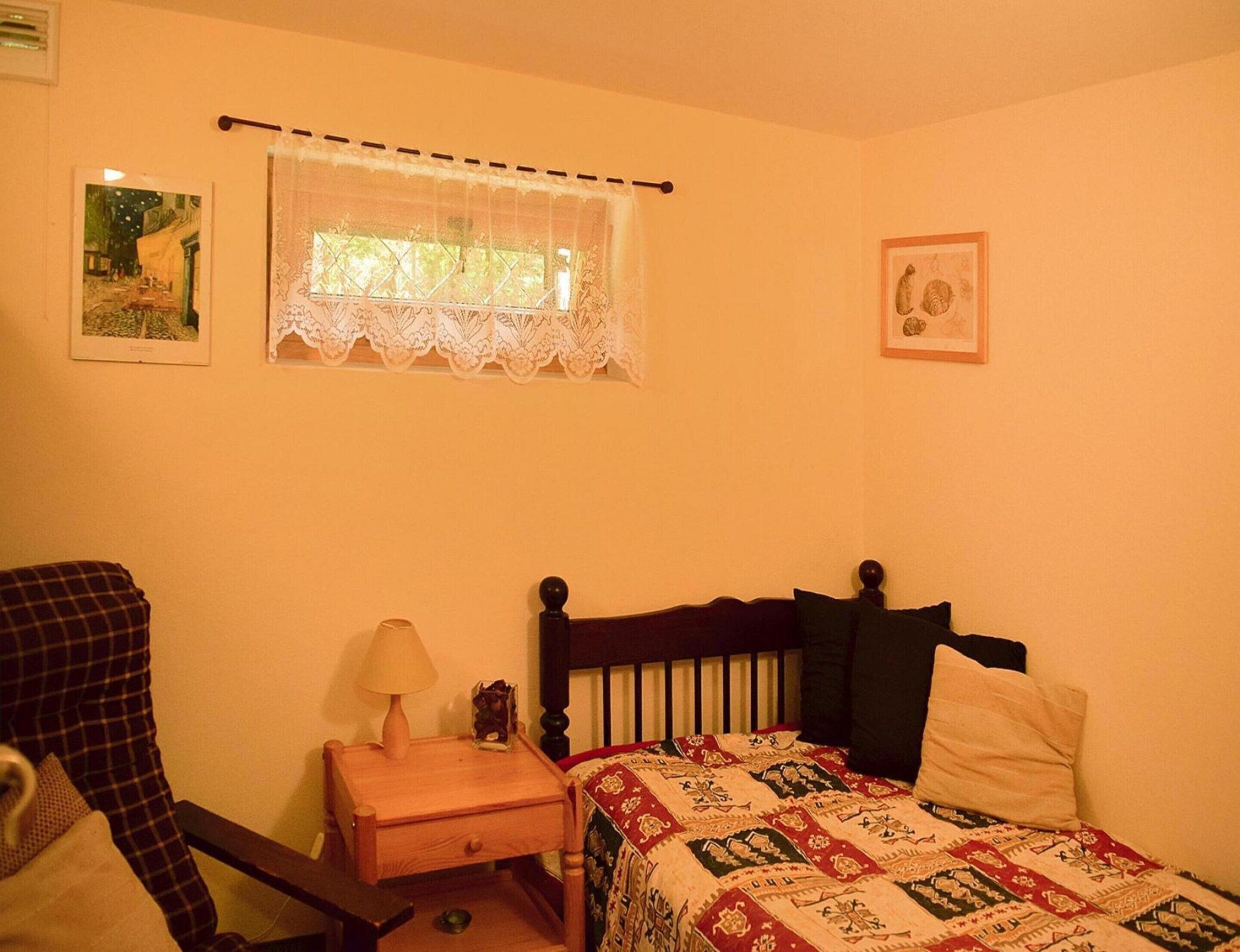 A bedroom with a wooden bed, a nightstand with a lamp, a window with lace curtains, and two framed pictures on the wall.