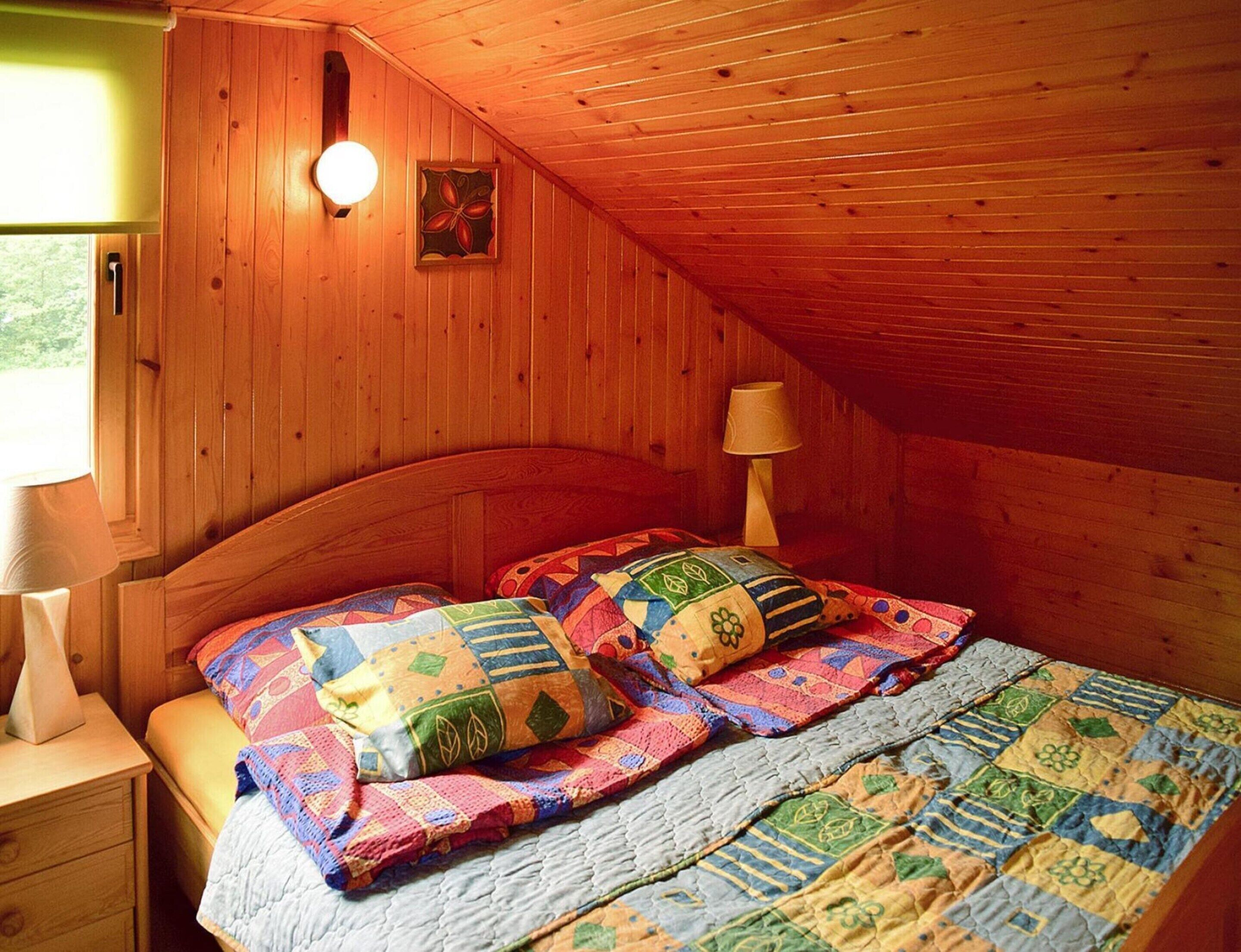 Une chambre confortable située sous les toits, avec des murs en bois, un plafond en pente, un lit recouvert d’une courtepointe colorée, une lampe de chevet et une fenêtre donnant sur un paysage verdoyant.