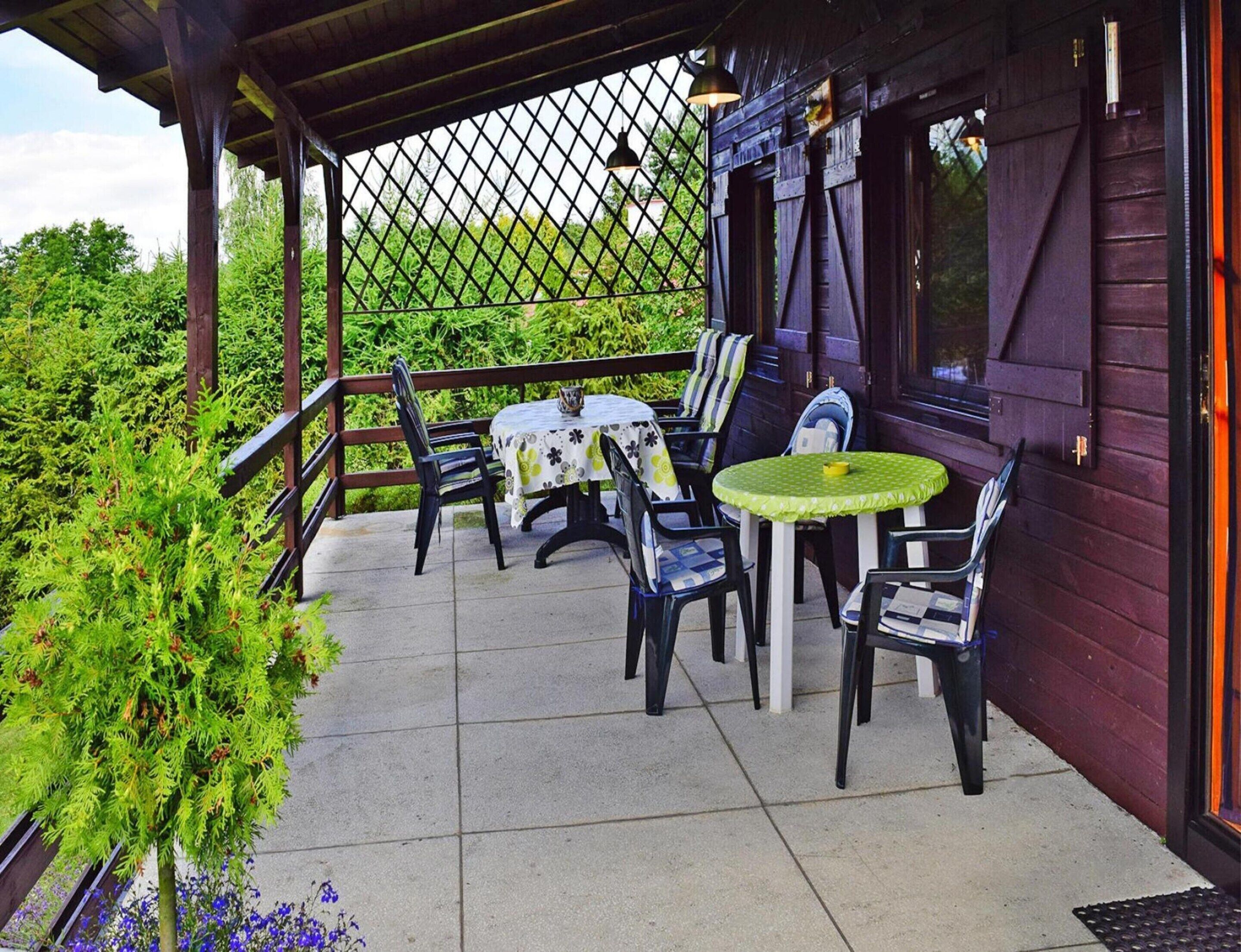 A covered outdoor seating area with tables and chairs, surrounded by greenery and a wooden fence.