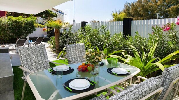 An outdoor dining area with a glass table, chairs, and a white fence in the background.
