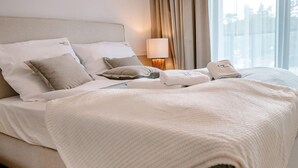 A neatly made bed with a white duvet and pillows, a bedside lamp, and a view of the outdoors through large windows. - Beautiful Apartment Very Close to the Beach (Rewal)