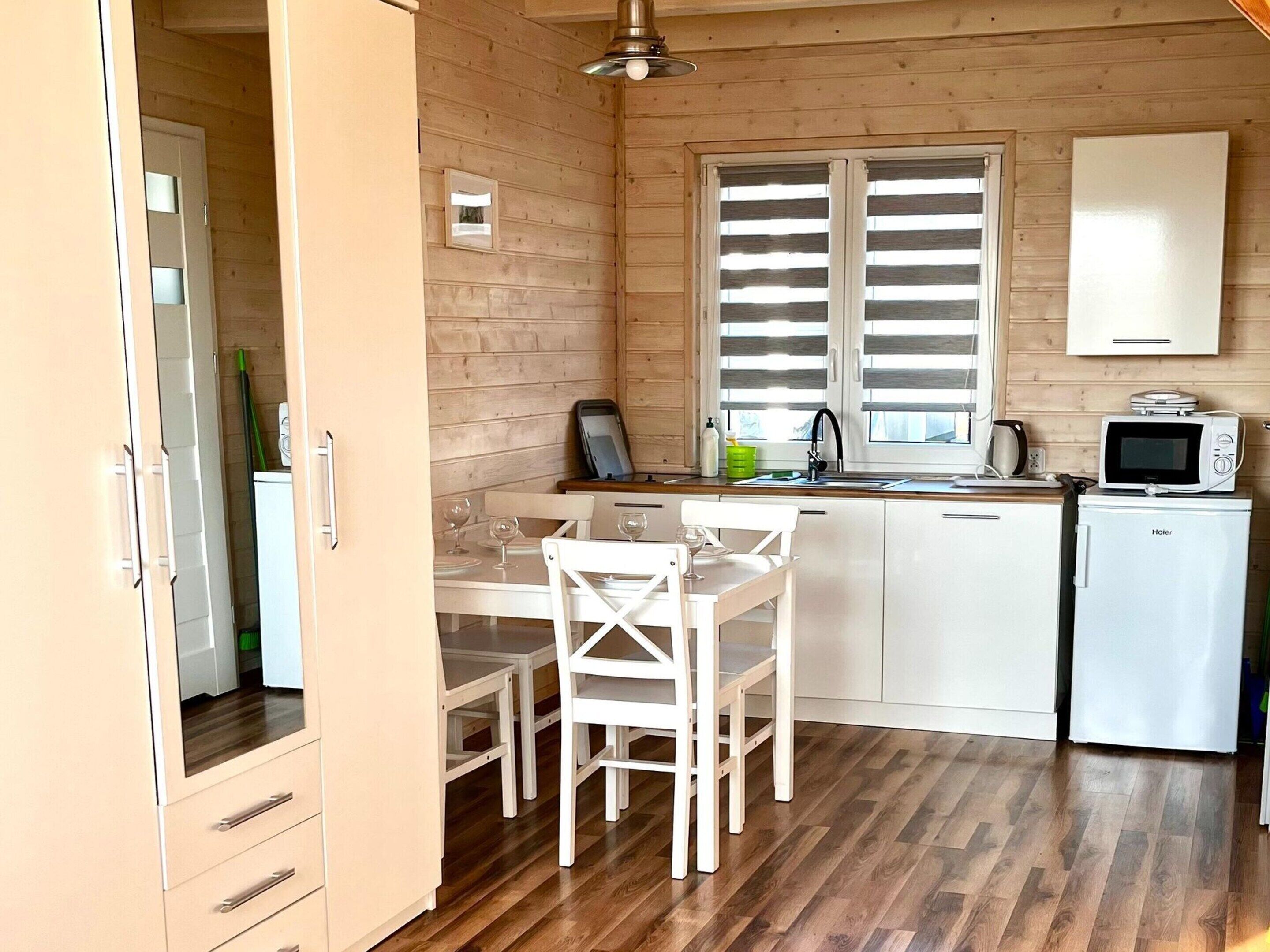 A compact kitchen with wooden walls, a dining area with white chairs, a small refrigerator, and a microwave.