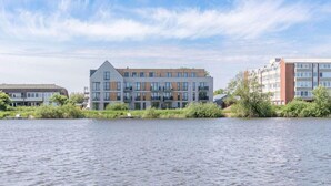 A modern multi-story building with a flat roof, large windows, and a mix of brick and glass facades, situated by a calm body of water. - Penthouse am Nordstrand auf dem Deich (Nordstrand)