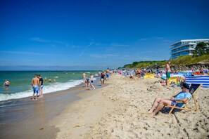 A beach with people sunbathing and swimming, a building in the background, and a clear blue sky. - Comfortable Apartment With a Terrace in Mielno (Mielno)
