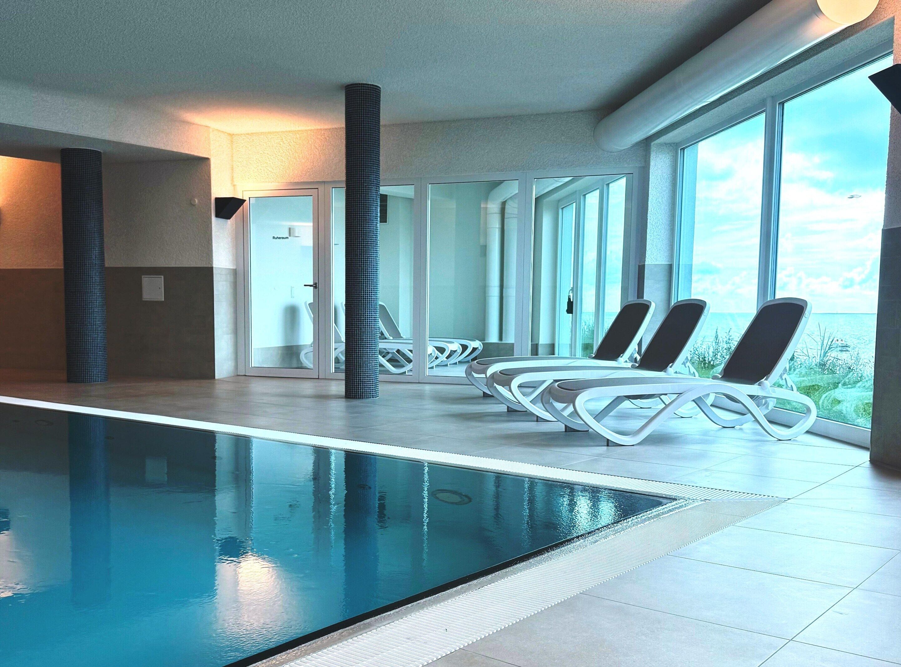 Un espace piscine intérieur moderne, doté de parois en verre transparent, de chaises longues et offrant une vue sur la mer.