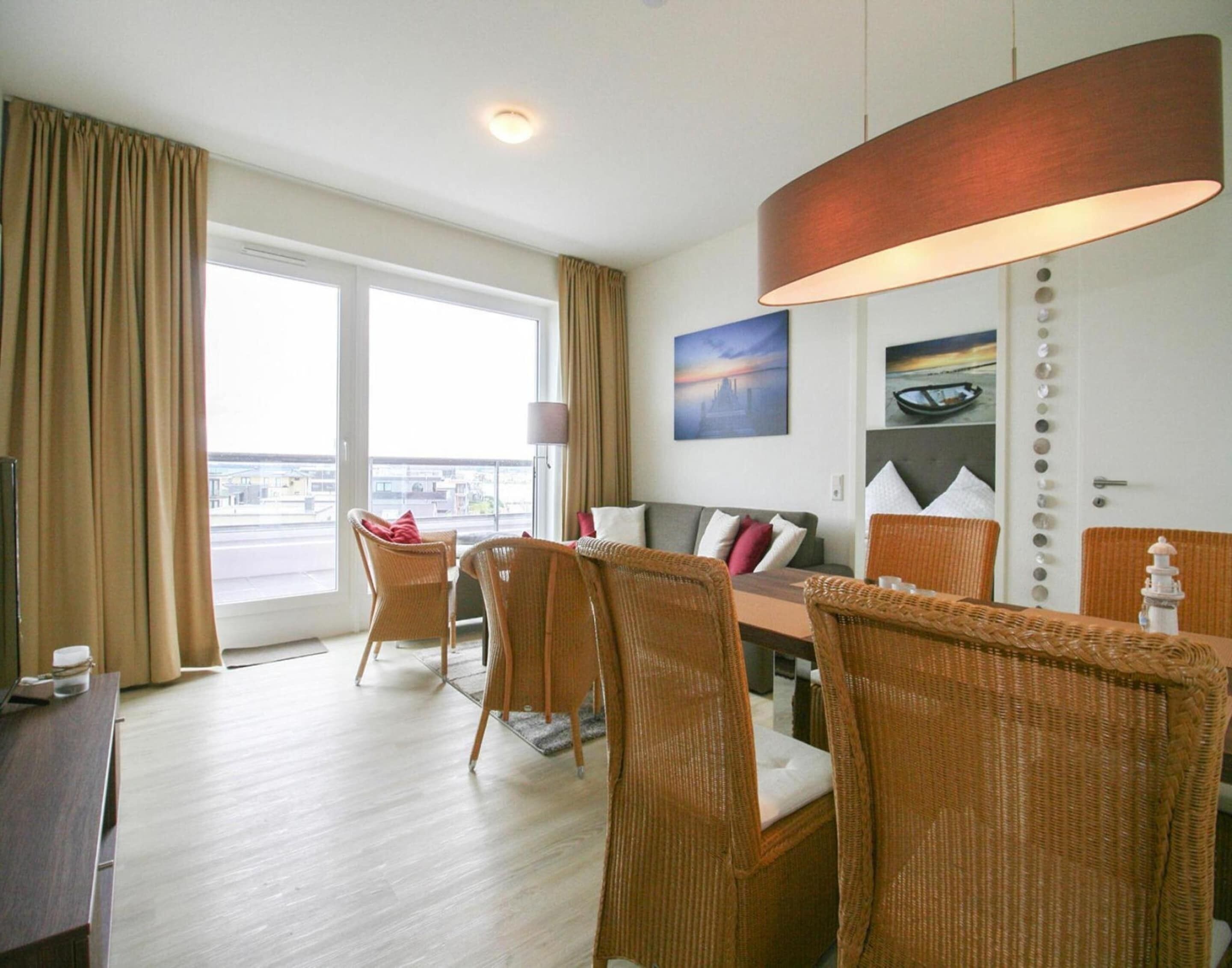 Ein Hotelzimmer mit Essbereich, Korbmöbeln, einem großen Fenster mit Vorhängen, einer Wandleuchte und einem Gemälde.