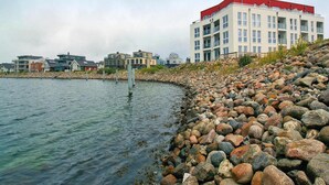 Una propiedad frente al agua con costa rocosa, un edificio de techo rojo y varias unidades residenciales al fondo.