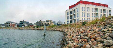 Um imóvel à beira-mar com costa rochosa, um edifício com telhado vermelho e vários edifícios residenciais ao fundo.