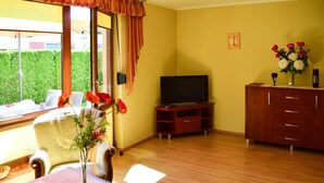 Un salon avec un plancher en bois, un téléviseur, un meuble en bois, un vase de fleurs et une fenêtre avec des rideaux.