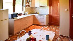 Une cuisine avec un sol carrelé, des placards en bois, un four à micro-ondes, un évier et une table recouverte d’une nappe à motifs floraux, avec un saladier rempli de fruits.