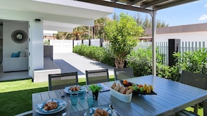 An outdoor dining area with a table set for breakfast, surrounded by chairs, and a view of a garden and a white building in the background.