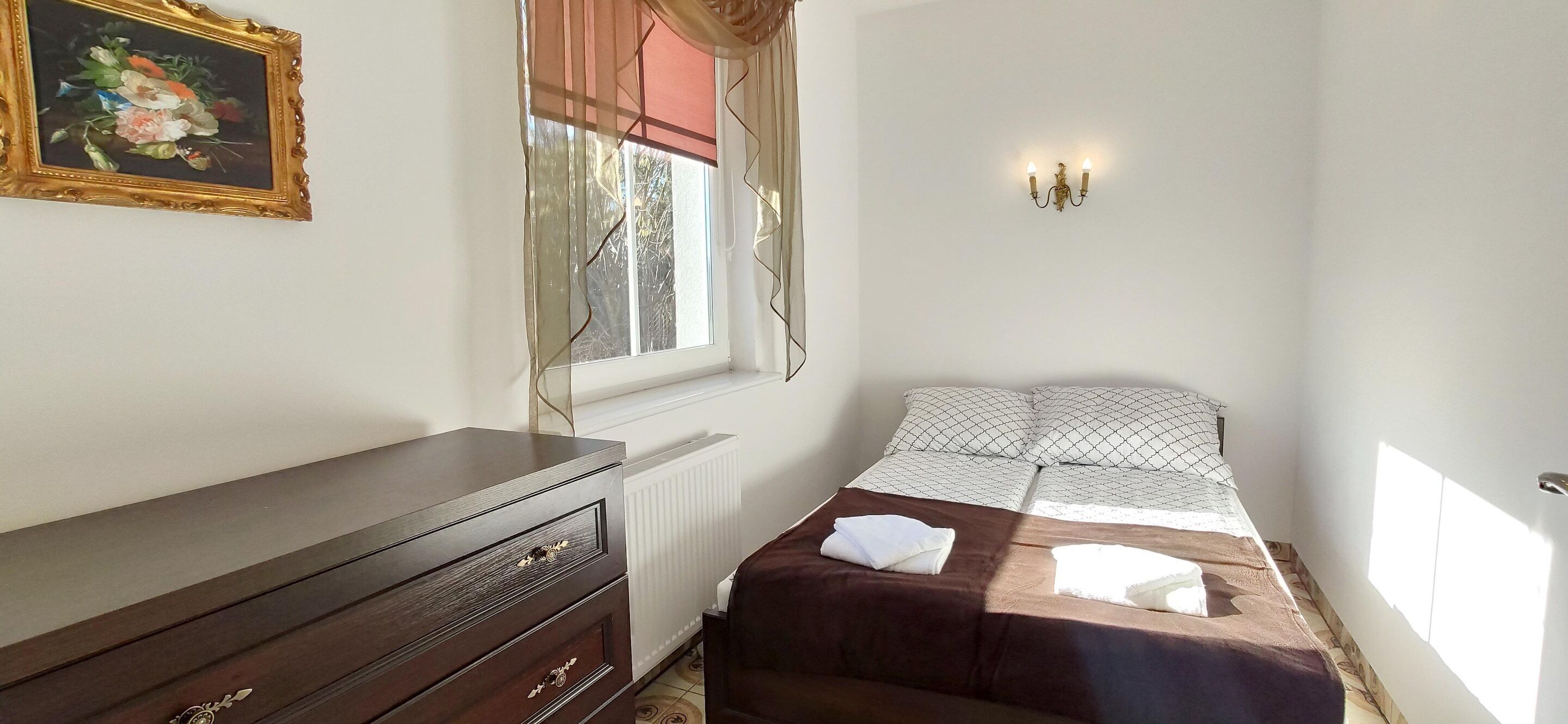 A bedroom with a bed, a dresser, a radiator, a window with curtains, and a wall-mounted light fixture.