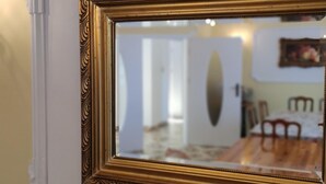 Un miroir doté d’un cadre doré orné, qui reflète une pièce comprenant un lit, une porte et un mur orné d’un tableau.