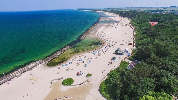 Vue aérienne d’une plage avec une longue étendue de sable, une digue rocheuse et une rangée d’éoliennes à l’horizon.