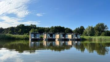 A row of floating houses with glass walls, situated on calm water, surrounded by greenery.