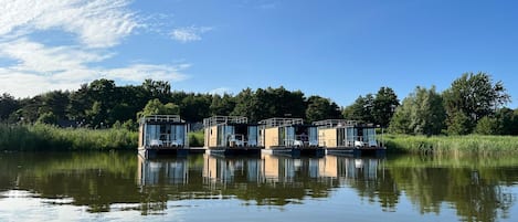 A row of floating houses with glass walls, situated on calm water, surrounded by greenery.