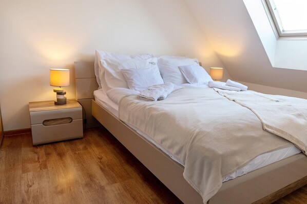 A neatly made bed with white linens and pillows, a bedside table with a lamp, and a wooden floor. - Comfortable Apartment Close to the Beach (Rewal)