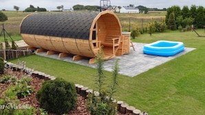 A wooden barrel-shaped structure with a ladder, a small table, and a blue inflatable pool in a grassy yard. - Holiday Home in Mielno Near Beach & Lake (Mielno)