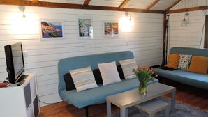 Ett mysigt vardagsrum med en blå soffa, ett litet bord och en tv på ett vitt skåp. Det hänger tre tavlor på väggen och en lampa står till höger.