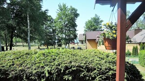 En välskött trädgård med tät buskvegetation, en hängande blomkruka och en byggnad med ett tegeltak i bakgrunden.