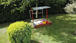 A small outdoor play area with a colorful playset, including a slide and a sandbox, surrounded by a neatly trimmed hedge and a tree. - Holiday Home in Kolczewo by the Lake (gmina Wolin)