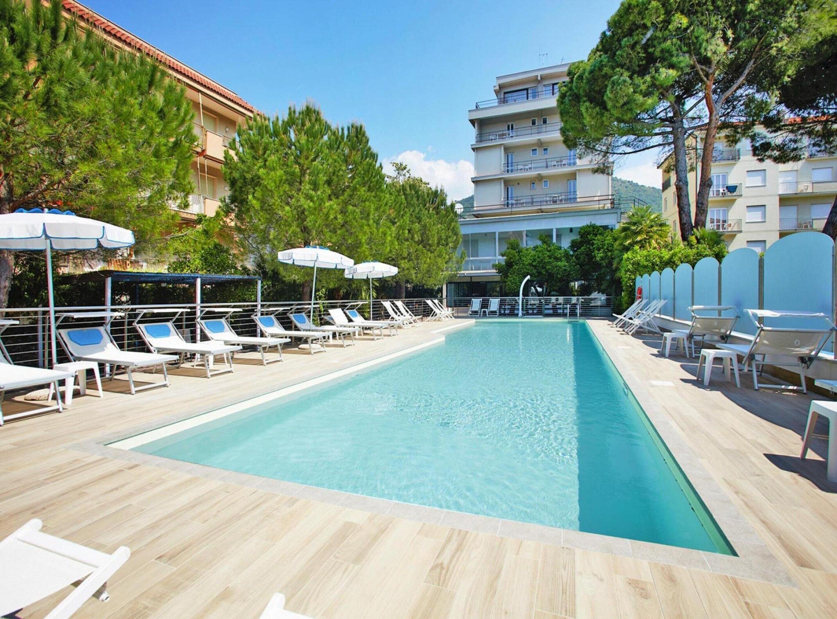 Une piscine entourée d’arbres et de bâtiments, avec des chaises longues et des parasols.