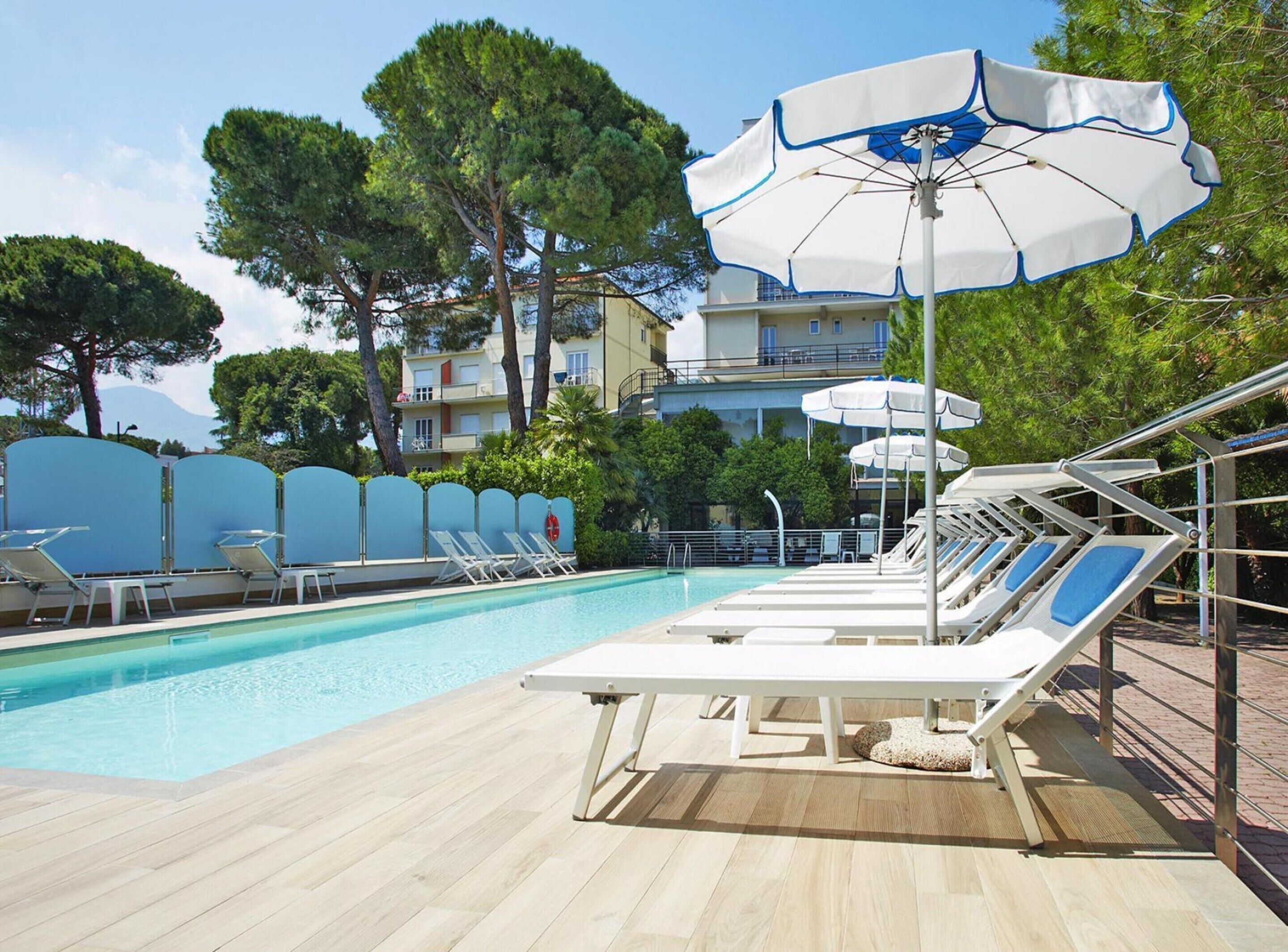 A poolside area with lounge chairs and umbrellas, surrounded by trees and a building in the background.