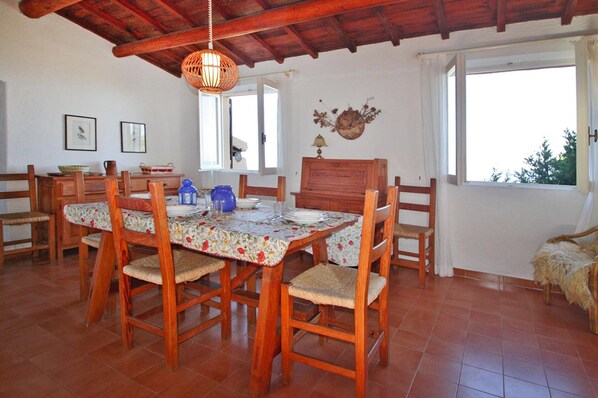 A dining area with a wooden table and chairs, a hanging light fixture, and a window with a view. - Apartment in Costa Paradiso With Garden Furniture (Trinità d'Agultu e Vignola)