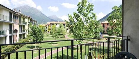 A balcony with a table set for dining, overlooking a courtyard with trees and a mountain in the background.