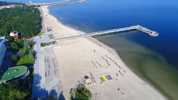 An aerial view of a coastal area with a beach, a long pier extending into the water, and a large open space with a colorful structure. - Cozy Holiday Apartment Close to the Beach (Kołobrzeg)